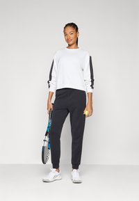 Λευκή μπλούζα με μαύρες ρίγες στα μανίκια, συνδυασμένη με σκούρες γκρι φόρμες. Το μοντέλο κρατάει μια ρακέτα τένις και μια μπάλα, φοράει λευκά αθλητικά παπούτσια.