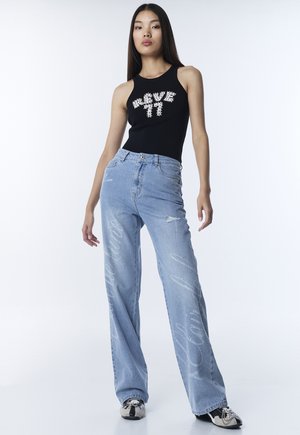 Jonge vrouw die een zwart mouwloos topje draagt met de tekst "REVE 77", lichtblauwe wijde jeans met witte letters en tweekleurige sneakers.