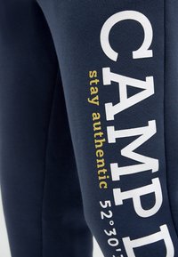 Marineblaue Hose mit weißem und gelbem Text, der auf dem Bein "CAMP stay authentic 52°30'39"" zeigt.