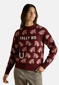 Frau in einem weinroten Pullover mit hellrosa Pferdeköpfen, dem Text "TALLY HO", einem Hufeisen und karierter Hose, steht mit einer Hand auf der Hüfte.