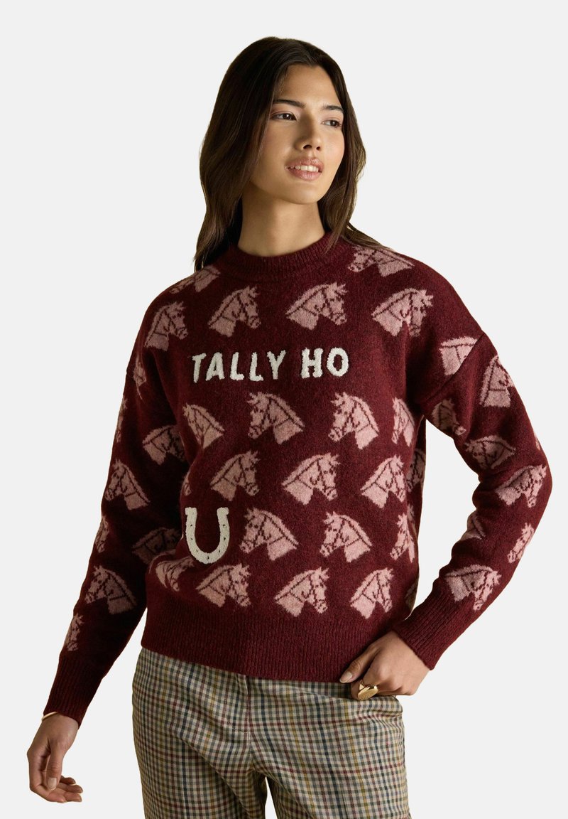 Frau in einem weinroten Pullover mit hellrosa Pferdeköpfen, dem Text "TALLY HO", einem Hufeisen und karierter Hose, steht mit einer Hand auf der Hüfte.