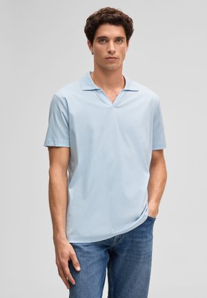 Junger Mann mit lockigem dunklem Haar, trägt ein hellblaues Polohemd mit kurzen Ärmeln und blaue Jeans, steht mit einer Hand in der Tasche.