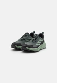 Pantofi de alergare pe potecă cu un superior verde închis, accente negre, talpă texturată și șireturi cu model. Vârf întărit și design ușor.