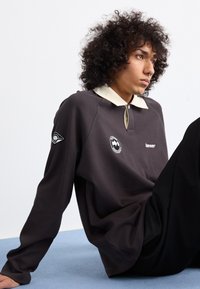 Mørkegrå sweatshirt med en beige krave, der har et broderet logo på brystet og et grafisk mærke på ærmet. Blød tekstur, løs pasform.