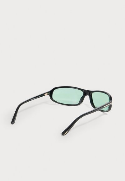 Lunettes de soleil rectangulaires noires avec des verres teintés vert clair placées sur une surface blanche, vues de dos et de côté.