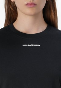 Artimas žmogaus kaklo nuotrauka, vilkinčio juodą marškinėlį su balta ženklu "KARL LAGERFELD".