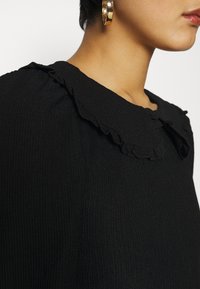 Svart ribbad topp med en volangkrage, i rund form. Närbild visar den strukturerade tyget och en subtil knappdetalj.