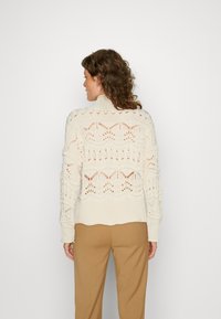 Witte gebreide trui met een opengewerkt design, voorzien van een hoge kraag en ribbel manchetten. Gecombineerd met beige broek.