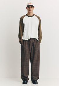 Cămașă albă cu mâneci lungi și maro, pantaloni largi maro în carouri și pantofi negri. Accesorii includ o șapcă albă cu text grafic roșu.