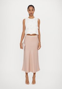 Weißes, kurzes Tanktop mit floralen Verzierungen, kombiniert mit einem hellrosa knielangen Rock, gehalten mit einer schlanken Clutch und nudefarbenen High Heels.
