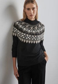 Schwarzer Wollpullover mit einem geometrischen Muster am Schulterbereich in Creme und Grau. Hat einen hohen Kragen und gerippte Bündchen und wird mit schwarzen Hosen kombiniert.