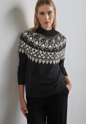 Schwarzer Wollpullover mit einem geometrischen Muster am Schulterbereich in Creme und Grau. Hat einen hohen Kragen und gerippte Bündchen und wird mit schwarzen Hosen kombiniert.