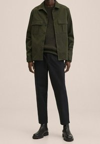 Giacca verde oliva con due tasche sul petto, chiusura a bottoni, maglione verde testurizzato sotto, abbinata a pantaloni neri a sigaretta e stivali neri.