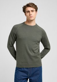Suéter de manga larga verde oliva con cuello redondo de canalé, tejido texturizado y mangas raglán, combinado con jeans de mezclilla azul.