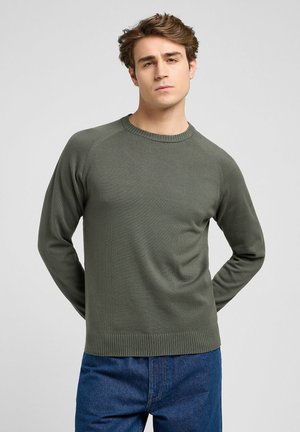 Pull olive en jersey à manches longues avec un col rond côtelé, en tissu tricoté texturé, et des manches raglan, assorti à un jean en denim bleu.