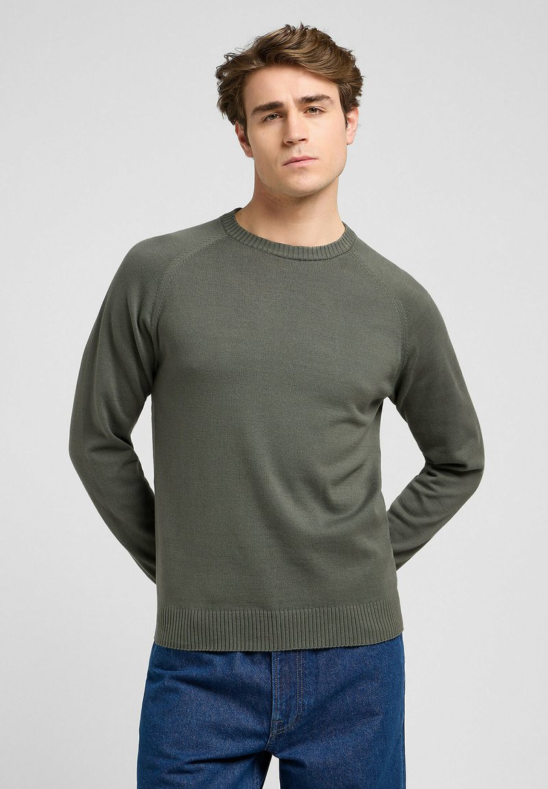 Suéter de manga larga verde oliva con cuello redondo de canalé, tejido texturizado y mangas raglán, combinado con jeans de mezclilla azul.
