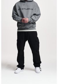 Sudadera con capucha gris con el texto "2Y STUDIOS", combinada con pantalones cargo negros con bolsillos y zapatillas blancas. Fondo liso.