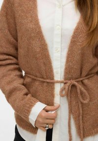 Žena nosi meku smeđu pletenu kardigan koju je zavezala u struku preko bijele košulje s dugim rukavima, držeći rub kardigana.