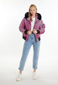 Giacca puffer con stampa leopardata rosa, colletto in pelliccia sintetica nera, chiusura con zip e polsini a coste, abbinata a jeans azzurri e sneakers bianche.