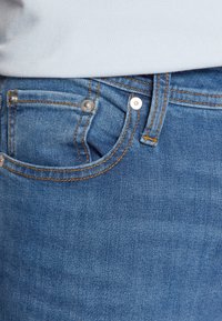 Blå denimjeans med kontrasterande orange sömmar, en framficka och metallnitar. Slät textur med en tight passform.