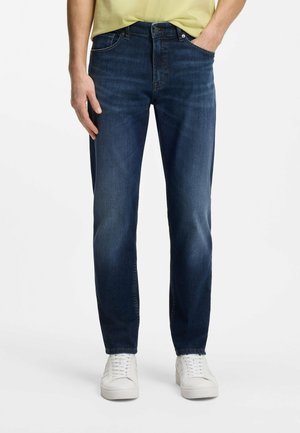 Uomo che indossa jeans aderenti blu scuro, sneakers bianche e una maglietta gialla, in piedi con una mano in tasca.