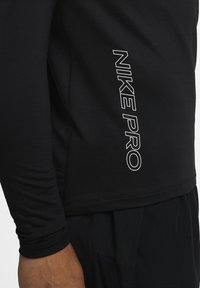 Nahaufnahme einer Person in einem schwarzen Langarmoberteil mit vertikal auf der Seite gedrucktem "NIKE PRO" und schwarzen Sportshorts.