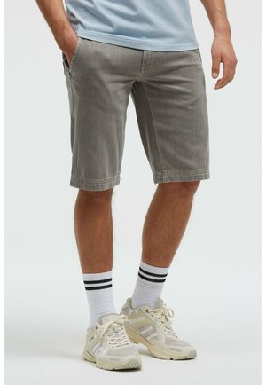Mann trägt graue knielange Shorts, weiße Socken mit schwarzen Streifen und hellfarbene Sportschuhe, steht vor einem schlichten Hintergrund.