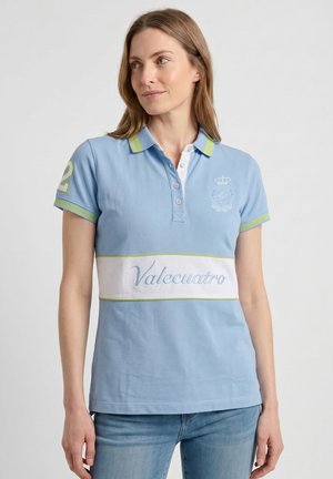 Mujer con un polo azul claro con ""Valecuatro"" en una banda blanca, ribete amarillo-verde y vaqueros azules, de pie contra un fondo liso.