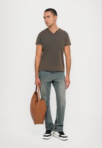Brown v-neck t-shirt, light blue jeans, black and white sneakers, holding a brown leather tote bag. Simple design and relaxed fit.
