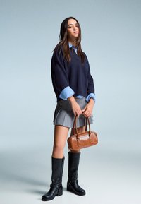 Mornarski oversized pulover, svijetloplava prugasta košulja, siva plisirana suknja, crne čizme do koljena s kopčama i smeđa kožna torba.