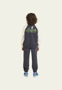 Tuta di un colore blu scuro con maniche crema, con il logo "Peanuts Sportswear" in verde. Sneakers blu con dettagli bianchi completano l'outfit.