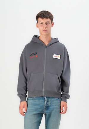 Gray zip-up hoodie with two front pockets, featuring red embroidered text and a logo patch on the chest. Soft fabric, relaxed fit.