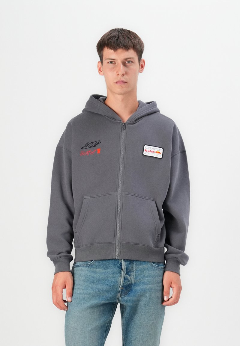 Gray zip-up hoodie with two front pockets, featuring red embroidered text and a logo patch on the chest. Soft fabric, relaxed fit.