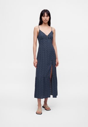 Kvinde iført en mørkeblå maxikjole uden ærmer med tynde stropper, lille hvidt blomsterprint, frontslids og beige flip-flop sandaler.