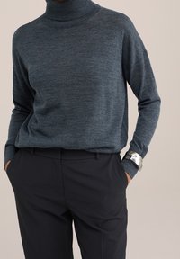 Pull à col roulé bleu foncé en tissu tricoté doux avec manches longues, associé à un pantalon noir sur mesure, agrémenté d'un élégant bracelet manchette en argent.