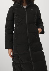 Kvinna iklädd en lång svart vadderad jacka med framsidans dragkedja, sidofickor och en liten vit CK-logotyp på bröstet.