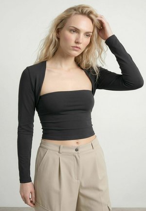 Lyshåret kvinde iført sort langærmet crop top og beige bukser med høj talje, rører ved håret og kigger let til siden mod en ensfarvet baggrund.