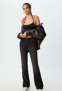 Giacca in ecopelle nera sopra un crop top nero, abbinata a jeans neri a vita alta e svasati. Il modello indossa sneaker argento e orecchini a cerchio.