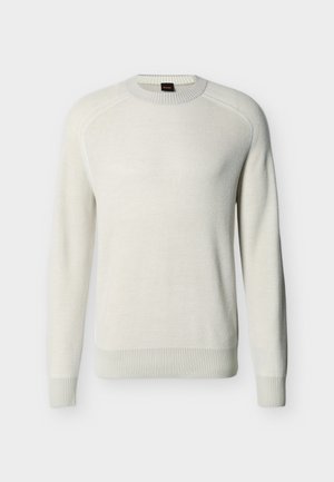 Gelsvai balta ilgomis rankovėmis megzta palaidinė su banguotais rankogaliais, apačia ir apykakle, ant vientisos spalvos fono.