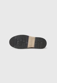 Schwarze Gummisohle mit strukturiertem Profil, versehen mit einem beigen Akzentetikett. Das Design umfasst verschiedene Grip-Muster für besseren Halt.
