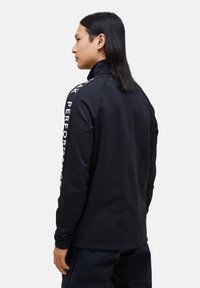 Sort zip-up jakke med højkrave og lange ærmer, med hvid tekst "PEAK PERFORMANCE" ned ad højre ærme og en glat tekstur.