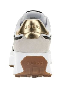 Sneaker bianco con accento in metallo dorato sul tallone, pannello in suede grigio, dettagli neri e suola in gomma marrone. Presenta il logo "GUESS".