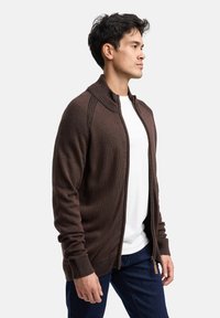 Brun strikket cardigan med lynlås, tekstureret krave, ribstrikkede ærmegab og en glat finish, lagt over en hvid t-shirt.