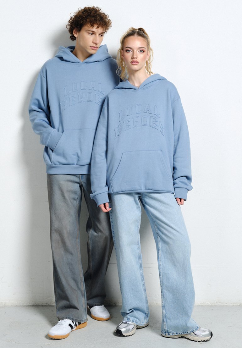 Młody mężczyzna i kobieta w dopasowanych jasnoniebieskich bluzach z kapturem z napisem „LOCAL HEROES”, noszący szerokie dżinsy i tenisówki, stojący na tle białej ściany.