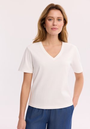 Kobieta z jasnobrązowymi włosami, ubrana w białą bluzkę na krótki rękaw z dekoltem w serek oraz niebieskie spodnie na jednolitym jasnym tle.