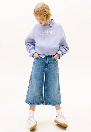 Jong persoon draagt een lavendelkleurige Tommy Hilfiger trui, blauwe wijde cropped jeans, witte sokken en witte sneakers, handen in de zakken.