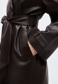 Σκούρο καφέ δερμάτινο παλτό τύπου τραντσκότ με ζώνη δεμένη στη μέση και χέρι στην μπροστινή τσέπη, ορατή λεπτομέρεια με λουράκι και κουμπί στο μανίκι.