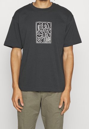 Person trägt ein schwarzes T-Shirt mit weißem abstraktem Textdesign auf der Brust und olivgrüner Hose, stehend vor einem schlichten weißen Hintergrund.