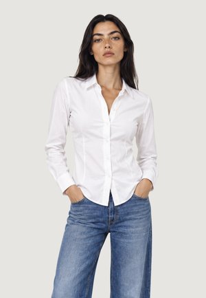 Femme portant une chemise blanche cintrée boutonnée et un jean bleu, les mains dans les poches, debout devant un fond clair uni.