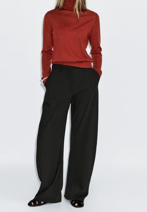 Woman wearing a rust-colored turtleneck sweater, black wide-leg trousers, black open-toe shoes, standing with hands in pockets.